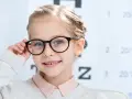 Uśmiechnięta dziewczynka w okularach, z tablicą optometryczną w tle. Okulary nosi się od jakiej wady, by widzieć wyraźnie.
