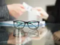 Okulary na pierwszym planie, w tle ręce przekazujące pieniądze. Idealny moment na skorzystanie z promocji na okulary!