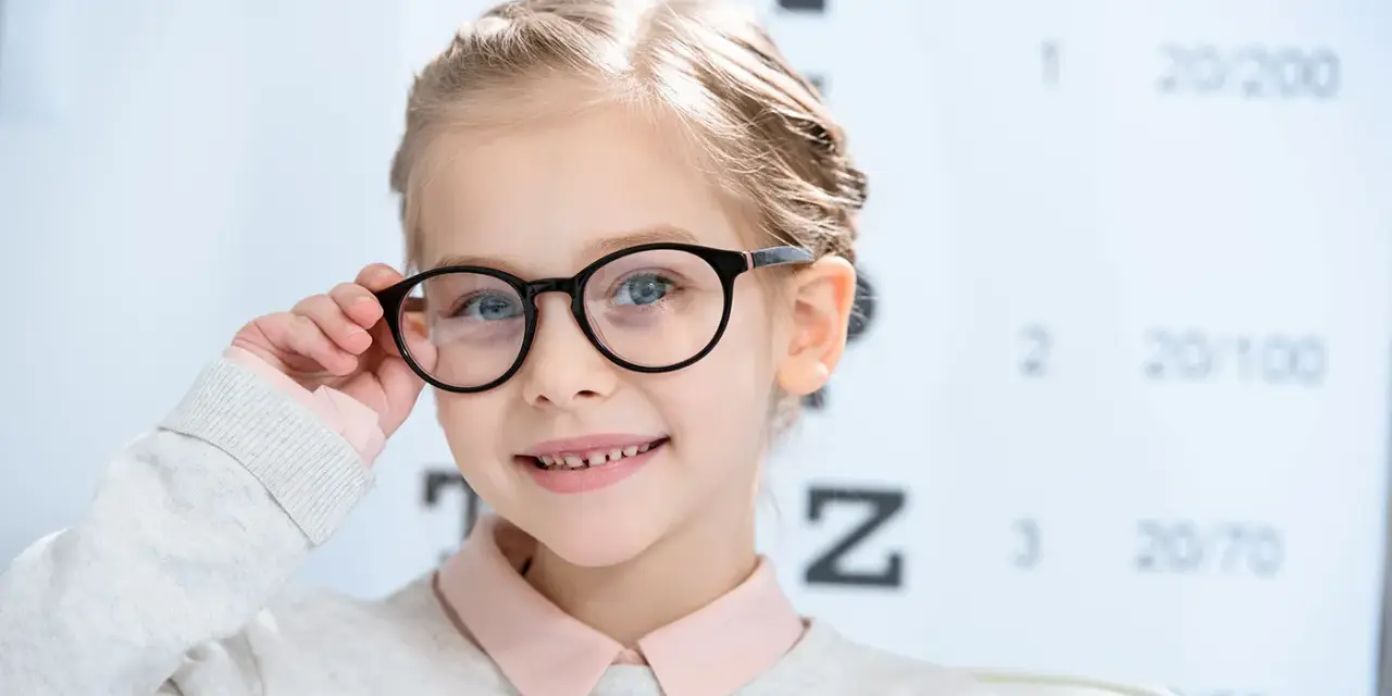 Uśmiechnięta dziewczynka w okularach, z tablicą optometryczną w tle. Okulary nosi się od jakiej wady, by widzieć wyraźnie.