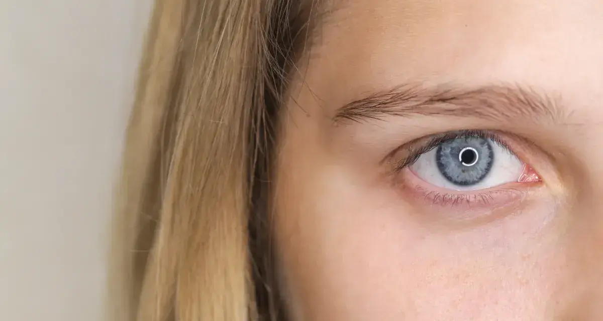Błękitne oko z jasną tęczówką, gdzie szpilkowate źrenice charakterystyczne są. Blond włosy opadają na twarz.