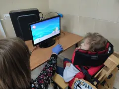 Tyflopedagog w niebieskiej rękawiczce pomaga dziecku w specjalnym foteliku grać w grę na komputerze.