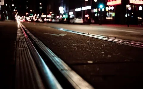 Nocne miasto, światła samochodów i neonów tworzą rozmyty obraz, jakby z powodu kurzej ślepoty. Trolejbusowe szyny błyszczą w ciemności.