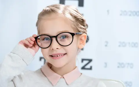 Uśmiechnięta dziewczynka w okularach, z tablicą optometryczną w tle. Okulary nosi się od jakiej wady, by widzieć wyraźnie.