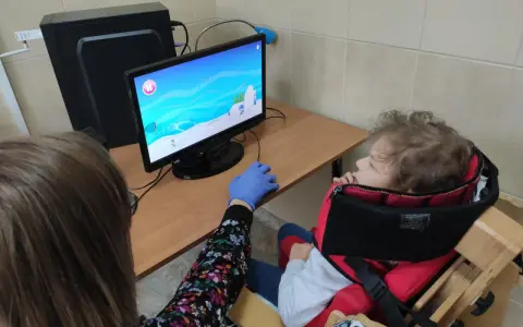 Tyflopedagog w niebieskiej rękawiczce pomaga dziecku w specjalnym foteliku grać w grę na komputerze.