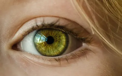 Zielona soczewka oka z refleksami światła, otoczona jasną skórą i blond włosami.