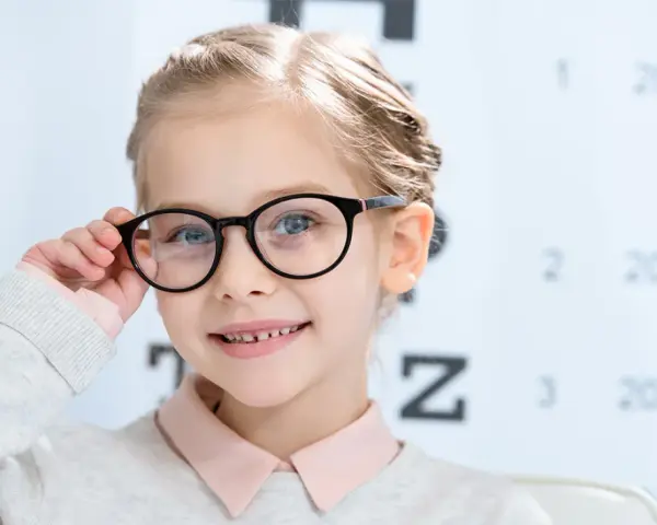 Uśmiechnięta dziewczynka w okularach, z tablicą optometryczną w tle. Okulary nosi się od jakiej wady, by widzieć wyraźnie.