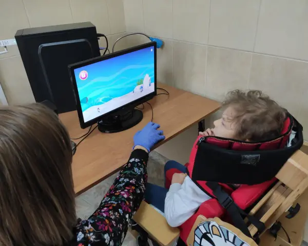 Tyflopedagog w niebieskiej rękawiczce pomaga dziecku w specjalnym foteliku grać w grę na komputerze.