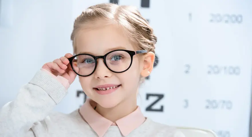 Uśmiechnięta dziewczynka w okularach, z tablicą optometryczną w tle. Okulary nosi się od jakiej wady, by widzieć wyraźnie.