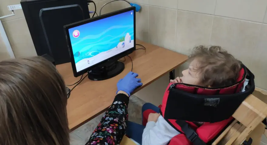Tyflopedagog w niebieskiej rękawiczce pomaga dziecku w specjalnym foteliku grać w grę na komputerze.
