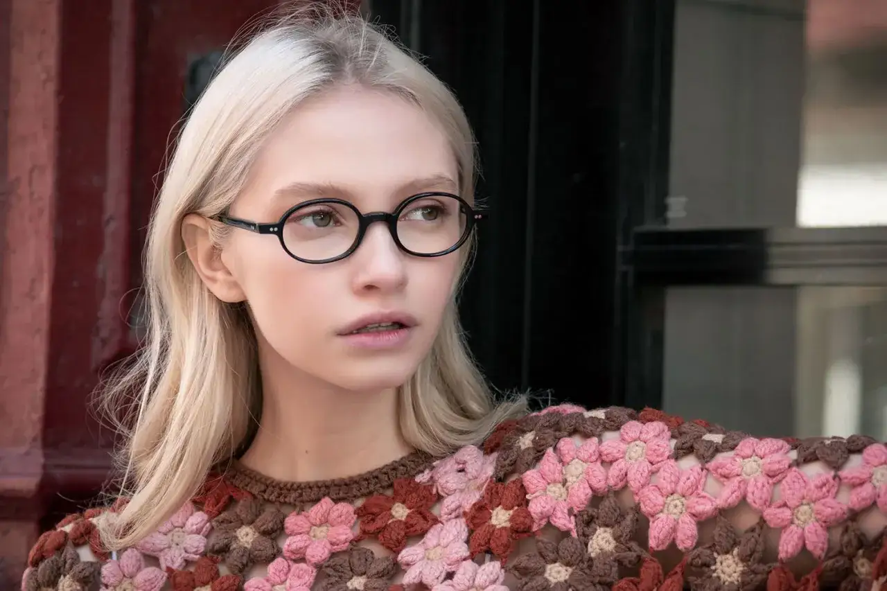 Blondynka w okrągłych, czarnych okularach, które są najmodniejsze oprawki do okularów. Ubrana w kolorowy, dziergany sweter w kwiaty.