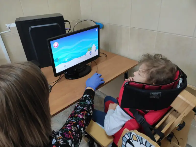 Tyflopedagog w niebieskiej rękawiczce pomaga dziecku w specjalnym foteliku grać w grę na komputerze.